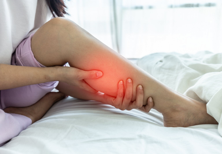 Femme assise sur un lit tenant son mollet douloureux avec une main, une rougeur localisée indique une tension musculaire. L’image illustre un massage ciblé des jambes et mollets proposé par le cabinet Terre de Shiatsu Lyon 5e, pour favoriser la détente musculaire et la circulation sanguine après l’effort.