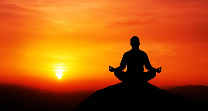 Personne assise les bras sur les genoux méditant à contre-jour au sommet d’une colline face à un coucher de soleil rouge-orangé, symbolisant le bien-être et la relaxation – cabinet Terre de Shiatsu Lyon 5e