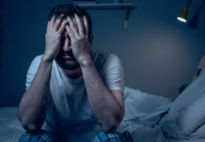 Homme assis sur un lit dans l’obscurité, le visage caché dans les mains, manifestement accablé par le stress ou l’anxiété nocturne. Cette image illustre les troubles du sommeil liés aux tensions mentales et met en avant l’efficacité d’un massage contre le stress au cabinet Terre de Shiatsu Lyon 5e pour favoriser un repos profond et apaisé.