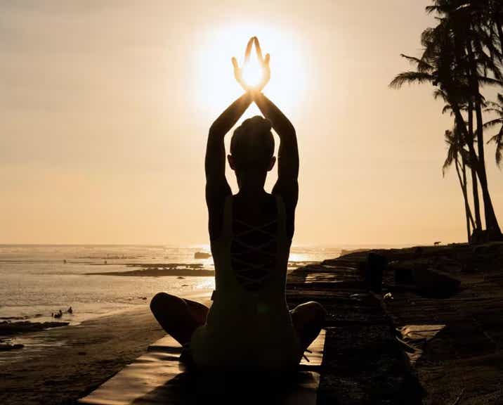 Femme en contre-jour assise jambes croisées sur une plage, de dos et face à l'océan, croisant ses mains au dessus de sa tête juste au milieu du soleil, symbolisant le bien-être général - cabinet Terre de Shiatsu Lyon 5e