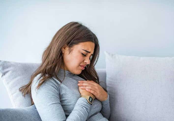 Femme assise sur un canapé, la main posée sur la poitrine, manifestant une gêne liée à un reflux acide. Cette image illustre le massage contre le reflux proposé par le cabinet Terre de Shiatsu Lyon 5e, visant à détendre la zone épigastrique, réduire le stress et améliorer le confort digestif.